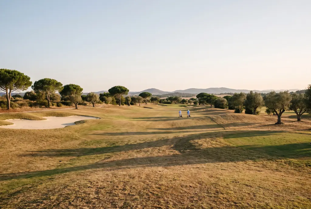Twee golfers wandelen met hun trolley over een zonnige fairway tussen pijnbomen en olijfbomen.